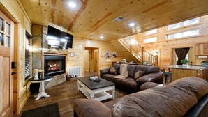 Un interior acogedor de cabaña de troncos con chimenea, sofás de cuero, un televisor y una escalera que lleva a un nivel superior.
