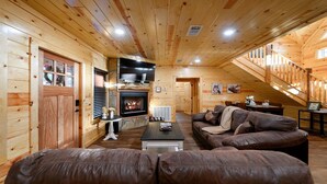 Interior acogedor de una cabaña de troncos con chimenea, sofás de cuero, televisor y una escalera que lleva a un nivel superior.