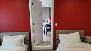 Habitación de hotel con dos camas, una pared roja y vistas a una zona de cocina a través de una puerta abierta.