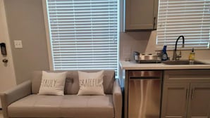 Una sala moderna con un sofá beige, dos almohadas con texto, una cocina con un lavaplatos y armarios, y un ventanal con persianas.
