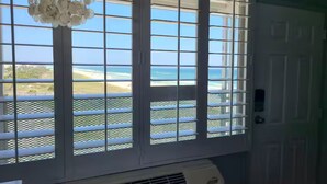 A room with a large window featuring white blinds, a view of the beach, and a white air conditioning unit below the window. - Beachfront Studio • Gulf Views • Pool • Walk to Beach + Dining (Panama City Beach)