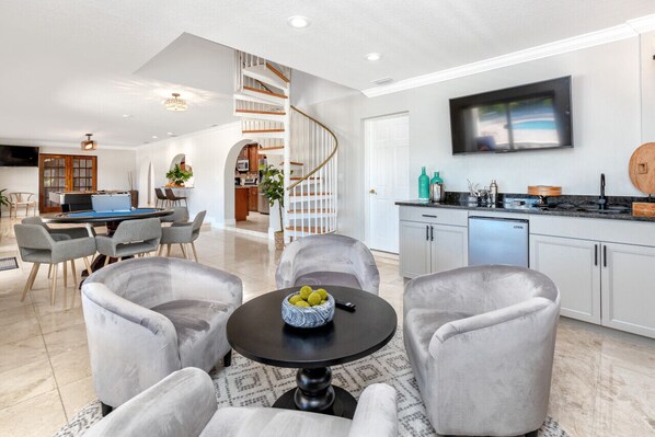 A modern kitchen with white cabinets, a black countertop, and a built-in oven. There is a dining area with grey upholstered chairs and a round table.