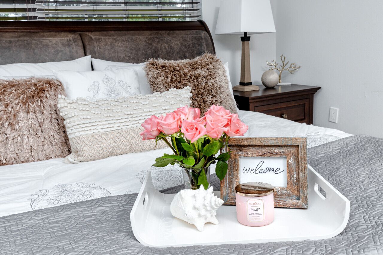 A neatly arranged bedroom with a bed, pillows, a bedside table, a lamp, a tray with a candle, a vase with flowers, and a welcome sign.