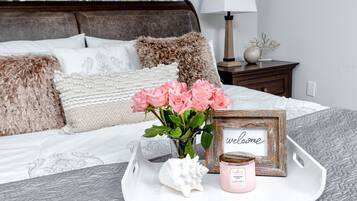 A neatly arranged bedroom with a bed, pillows, a bedside table, a lamp, a tray with a candle, a vase with flowers, and a welcome sign.