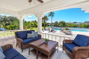 Un espace extérieur abrité, aménagé avec des meubles en osier, une piscine et offrant une vue sur un lac et des palmiers.
