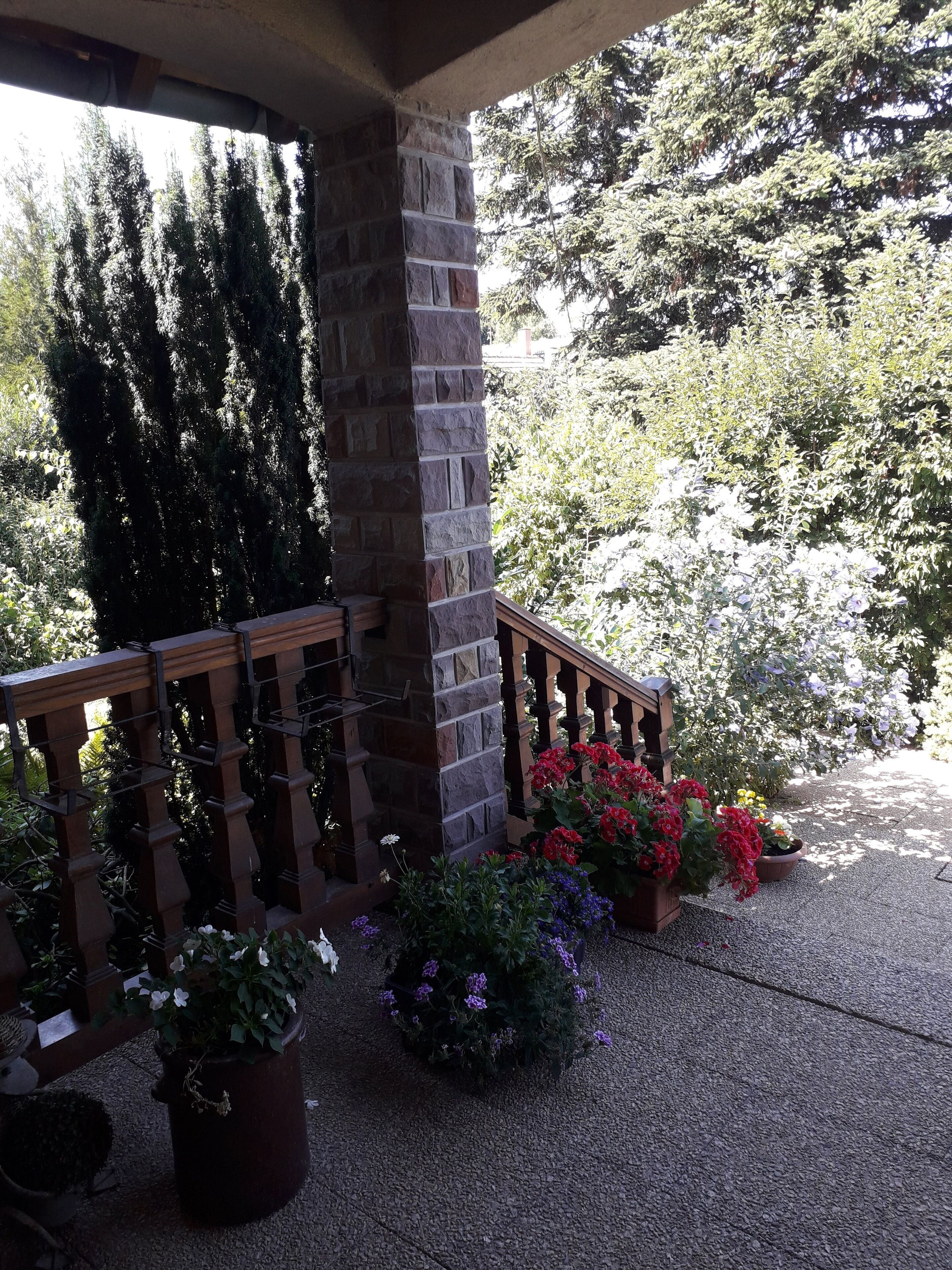 A covered outdoor area with a stone pillar, wooden railing, and potted plants with flowers.