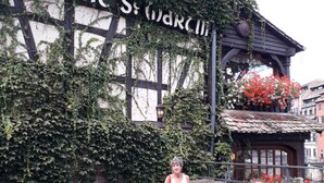 Une femme est assise à une table à l’extérieur, entourée de chaises et d’un banc en bois, et un panneau indique : « La PONC St. Marcill ».