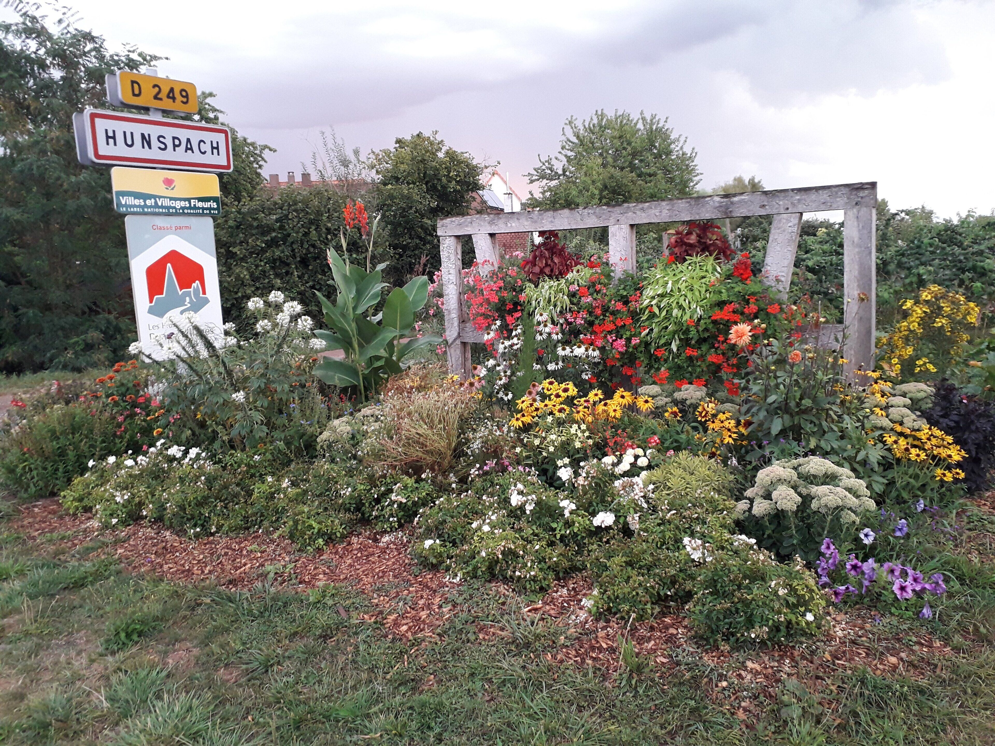 Un cartel con 'HUNSPACH' y 'D 249' señalando una ubicación, rodeado de una variedad de flores y plantas coloridas.