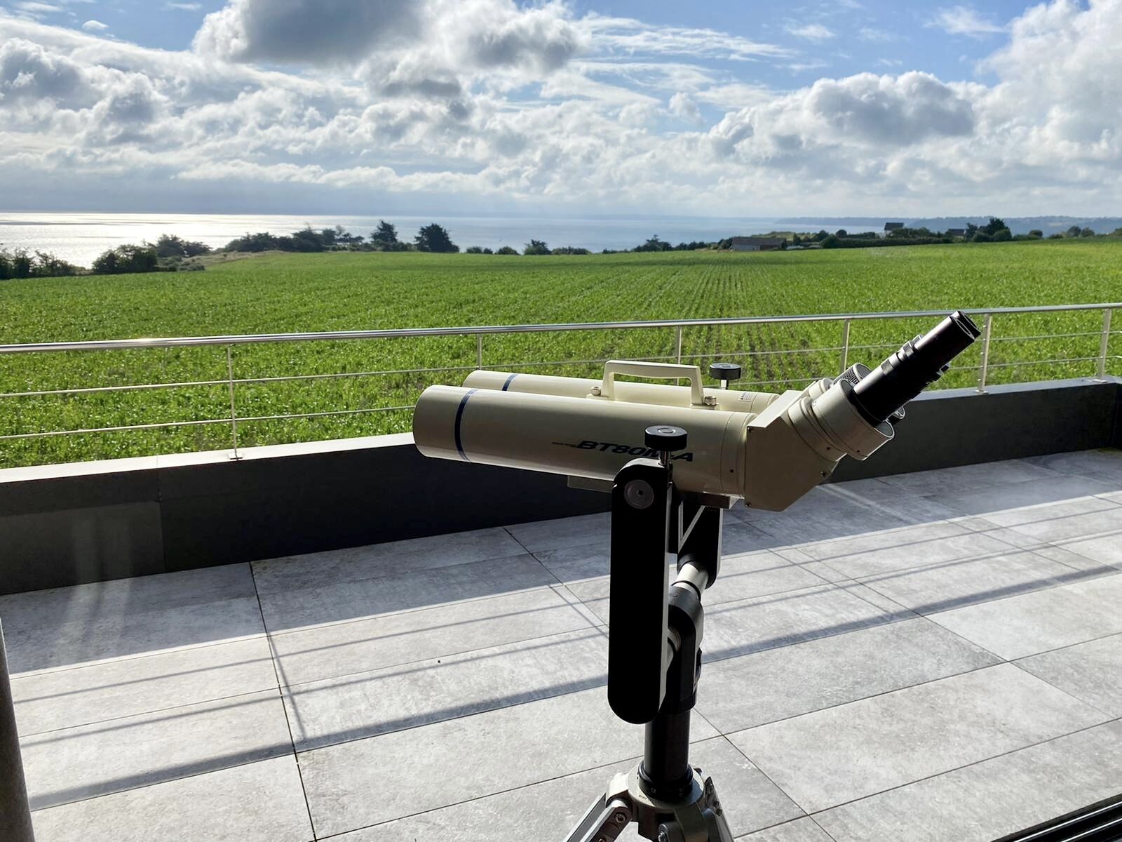 Un télescope sur un balcon, avec vue sur un champ verdoyant et la mer.