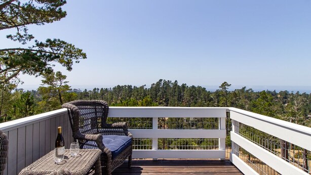 Una terraza de madera con mobiliario de mimbre y barandilla blanca, con vistas a un paisaje boscoso.
