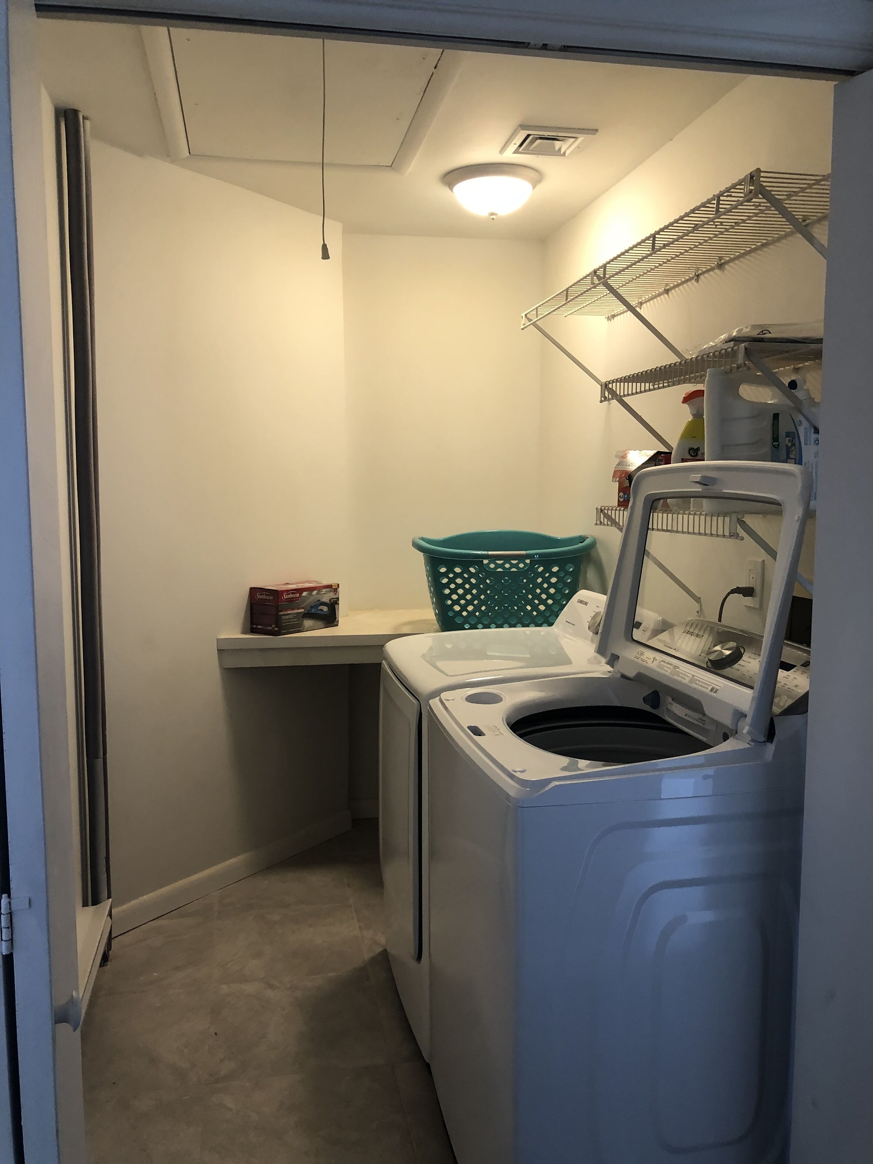 A compact laundry room with a front-loading washing machine, a drying rack, a shelf with cleaning supplies, and a green laundry basket.