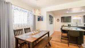 Un comedor con una mesa de madera, bancos y un camino de mesa estampado. Hay una ventana con cortina, una lámpara colgante y obras de arte en las paredes.