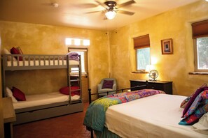 A room with bunk beds, a desk, a chair, a bed with a colorful blanket, a ceiling fan, and a window with blinds.