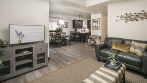 Ein modernes Wohnzimmer mit Sofa, Fernseher und Unterhaltungseinheit. Es gibt einen Essbereich mit Tisch und Stühlen, und im Hintergrund eine Küche.
