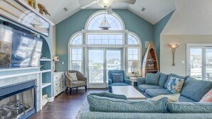 A spacious living room with a high ceiling, a fireplace, a large window, and a cozy seating area. - Corolla Coastin | 740 ft to the beach | Private Pool, Hot Tub (Corolla)