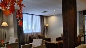 Un hall d’hôtel moderne avec un espace salon comprenant des fauteuils et un canapé, une grande colonne et une vue sur l’extérieur par une fenêtre agrémentée de rideaux.