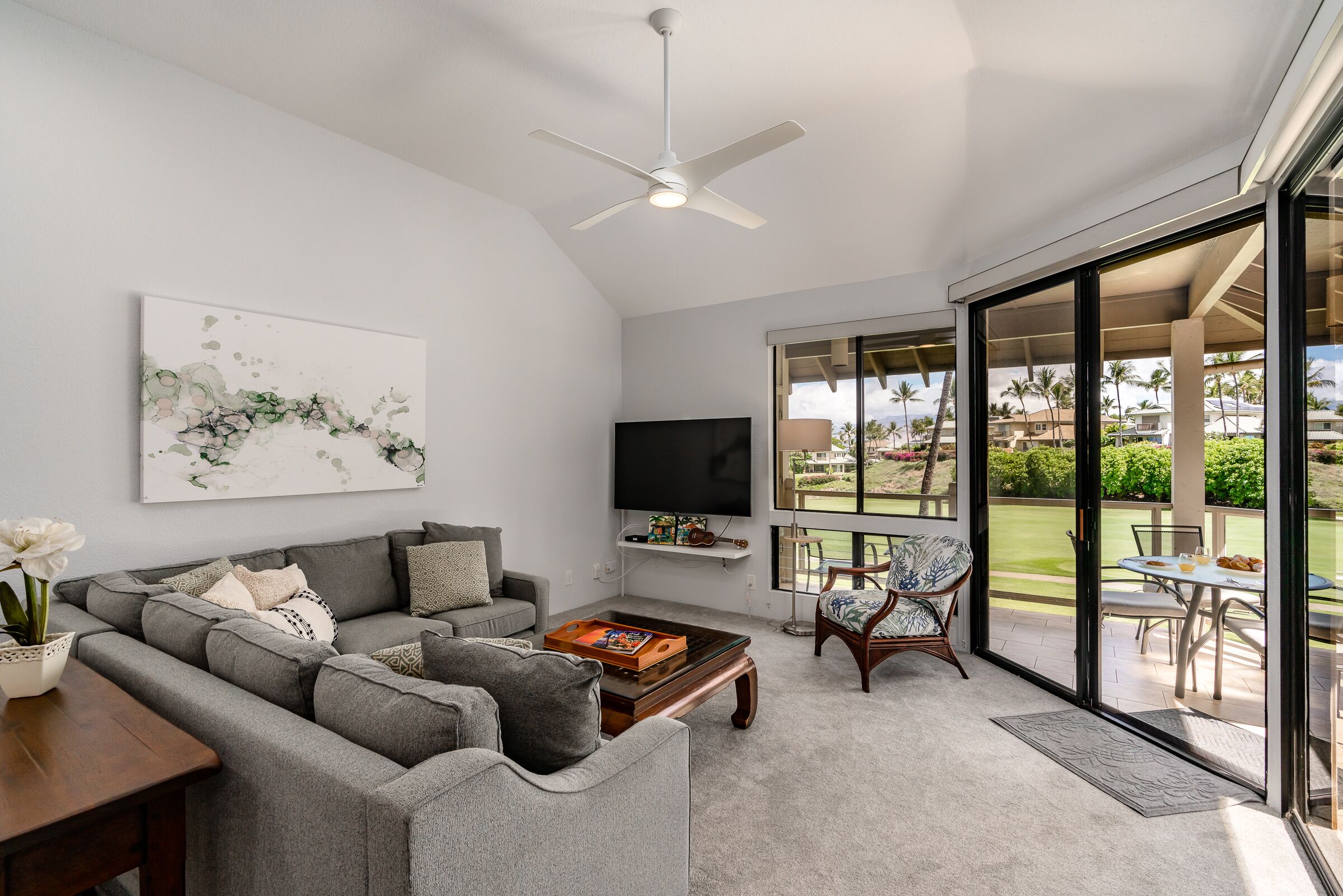 A modern living room with a grey sectional sofa, a coffee table, a flat-screen TV, and a large window offering a view of a garden and palm trees.