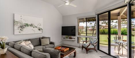 A modern living room with a grey sectional sofa, a coffee table, a flat-screen TV, and a large window offering a view of a garden and palm trees.