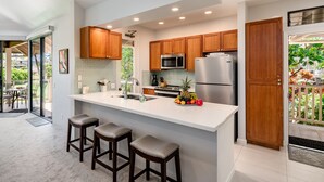 A modern kitchen with wooden cabinets, stainless steel appliances, a large island with stools, and a view of the outdoors. - Grand Champions in Wailea – Spacious Villa Near Pool w/ Ocean & Sunset Views (Wailea)