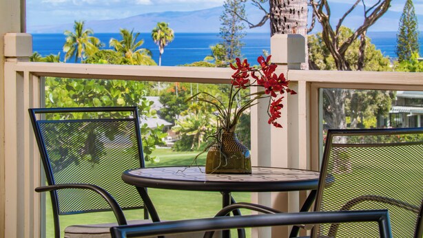 A balcony with a table and chairs, overlooking a tropical landscape with palm trees and a view of the ocean. - Grand Champions in Wailea – Spacious Villa Near Pool w/ Ocean & Sunset Views (Wailea)
