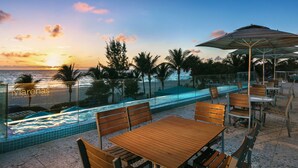 A poolside area with wooden tables and chairs, a glass barrier, and a view of the ocean at sunset.