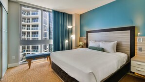 A hotel room with a large bed, a bench, a nightstand, a lamp, and a view of a high-rise building.