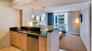 A modern kitchen with a glass island, wooden cabinets, and a dishwasher. There is a dining area with a table and chairs, and a large window offering a view of the city.