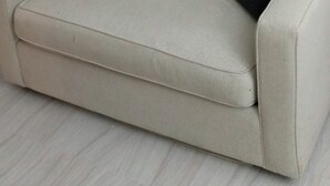 Un sofá color beige con dos cojines negros, un cuadro abstracto de colores en la pared y un radiador blanco.