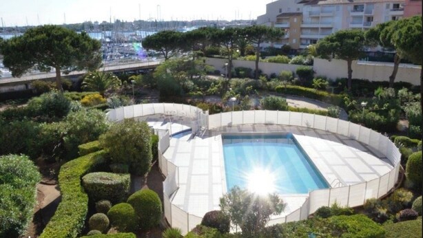 Ein kreisförmiger Pool mit einem sonnendurchfluteten Zentrum, umgeben von gepflegten Büschen und Bäumen, mit einem Yachthafen und Gebäuden im Hintergrund.
