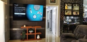 A modern living room with a flat-screen TV mounted on the wall, a wooden console table with decorative items, a gray sofa, and a kitchen area with a tiled backsplash and a fish tank.