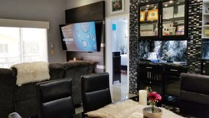 Un salon moderne avec un canapé gris, des chaises noires, un téléviseur et un coin cuisine doté d’un mur recouvert de carreaux.