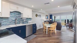 A modern kitchen with a marble countertop, blue cabinets, and a backsplash. There is a dining area with a table and chairs, and a view of the ocean through large windows.