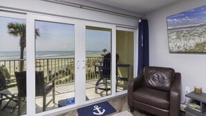 A balcony with a view of the beach, a chair, and a table.