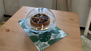 Un tavolo in legno con una torta di vetro su un sottopiatti a motivi fogliari, un asciugamano appeso al muro e una finestra con vista sul mare.