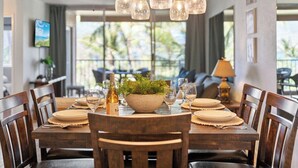 A dining area with a wooden table set for four, a bowl of green plants, and wine glasses. - Remodeled BeachFront 2BD Whale Watching-Menehune Shores 402 (Kihei)