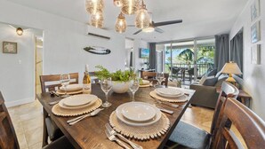 A dining area with a wooden table set for four, surrounded by chairs. There are decorative lights above the table, and a ceiling fan is visible. - Remodeled BeachFront 2BD Whale Watching-Menehune Shores 402 (Kihei)