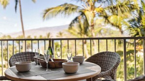 A round table with four bowls and three wine glasses, set on a balcony with wicker chairs and a view of palm trees and a mountain. - Remodeled BeachFront 2BD Whale Watching-Menehune Shores 402 (Kihei)