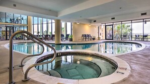 A large indoor swimming pool with a curved design, surrounded by lounge chairs and a glass-enclosed area with a view of palm trees. - Dauphin Island Condo w/ Pool, Balcony & Ocean View (Dauphin Island)