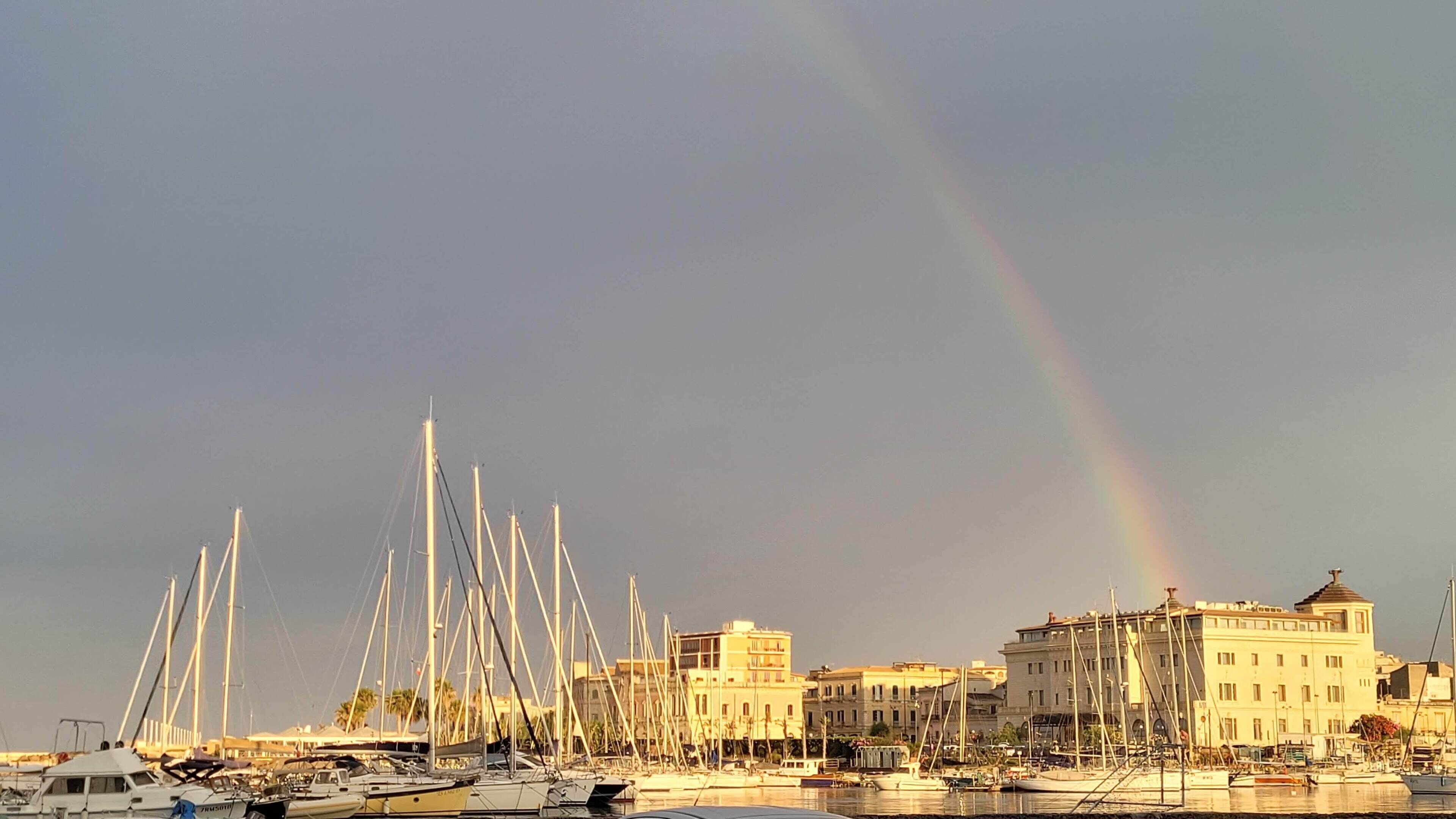 A marina with numerous sailboats docked, a rainbow in the sky, and buildings in the background.