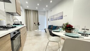 A modern kitchen with a gas stove, microwave, and wooden cabinets. There is a dining area with a round table, chairs, and a vase with flowers. - Appartamento Camera da Letto Vista Mare, 2 Bagni Parcheggio,spiaggia Solarium (Siracusa)