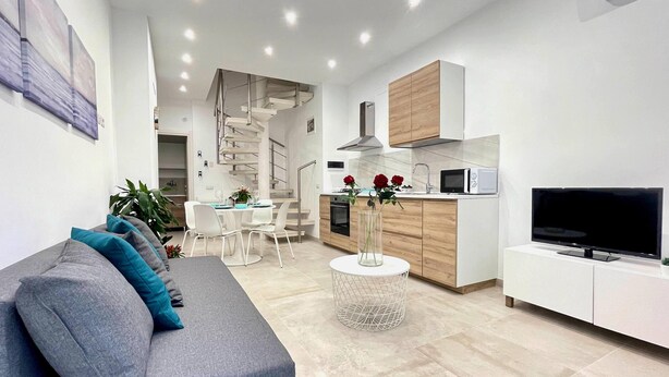 A modern living room with a grey sofa, a dining area with a white table, and a kitchen with wooden cabinets and a TV. - Appartamento Camera da Letto Vista Mare, 2 Bagni Parcheggio,spiaggia Solarium (Siracusa)