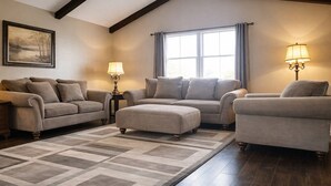 A living room with a sectional sofa, two armchairs, a coffee table, and a rug. There are two lamps on side tables, a painting on the wall, and a window with curtains. - The Illini Game House (Champaign)