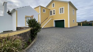 A two-story yellow building with a green garage door, a white exterior wall, and a paved driveway. - Casa do Caminho (Praia da Vitoria)