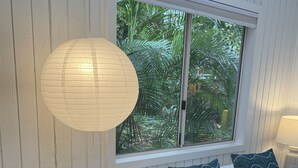 A bedroom with a bed, white bedding, a window with a view of greenery, a bedside table with a lamp, and a decorative paper lantern. - Sun Filled, Tropical Bungalow (Haleiwa)