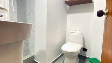 A bathroom with a toilet, a shower area, a sink, and a green trash bin.