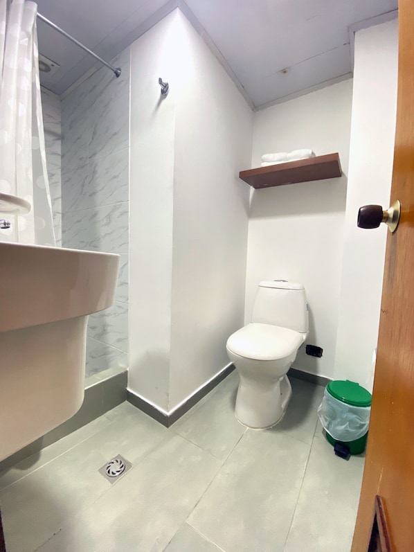 Un baño con inodoro, zona de ducha, lavamanos y un cubo de basura verde.