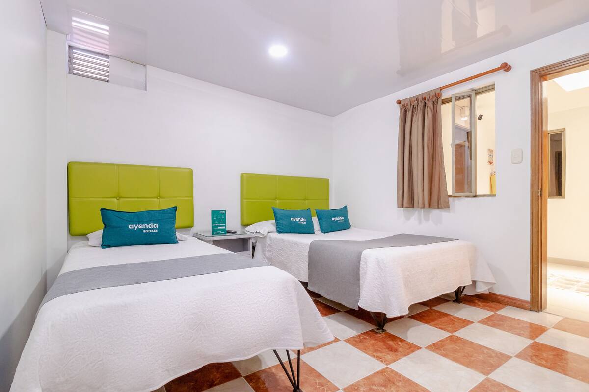 Habitación de hotel con dos camas, cada una con cabecero verde y almohadas turquesa, una mesita de noche pequeña y un quiosco de registro.