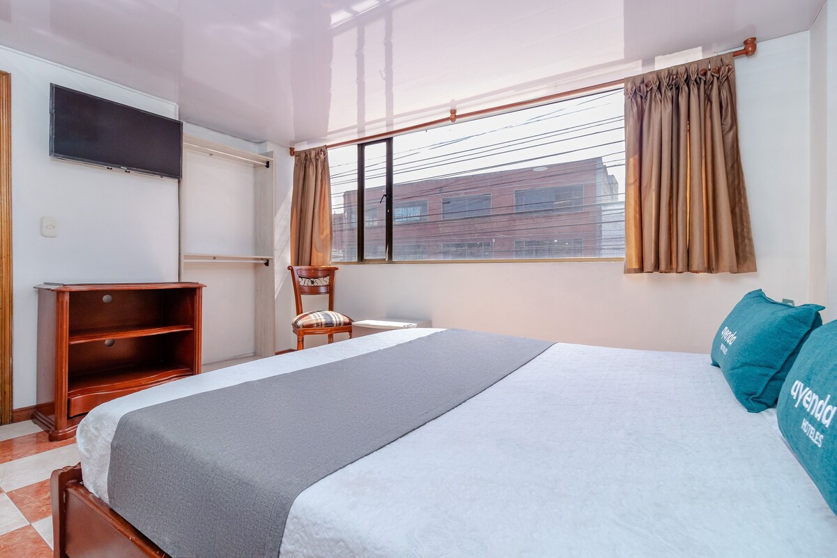Ein Hotelzimmer mit Bett, Fernseher, Fenster mit Vorhängen, einem Sessel und einem Holzregal.