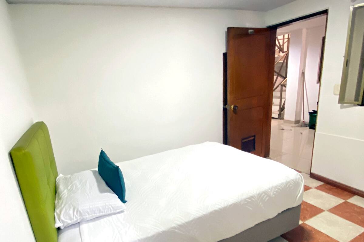 Una cama individual con cabecera verde y ropa de cama blanca, una mesita de noche pequeña y suelo de baldosas.