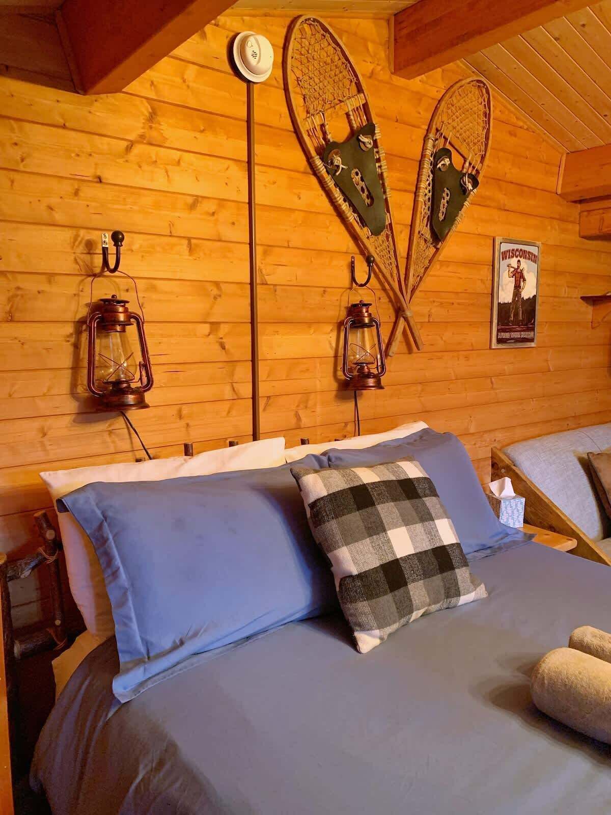 A wooden cabin room with a bed, two lantern-style wall lights, and a poster on the wall.